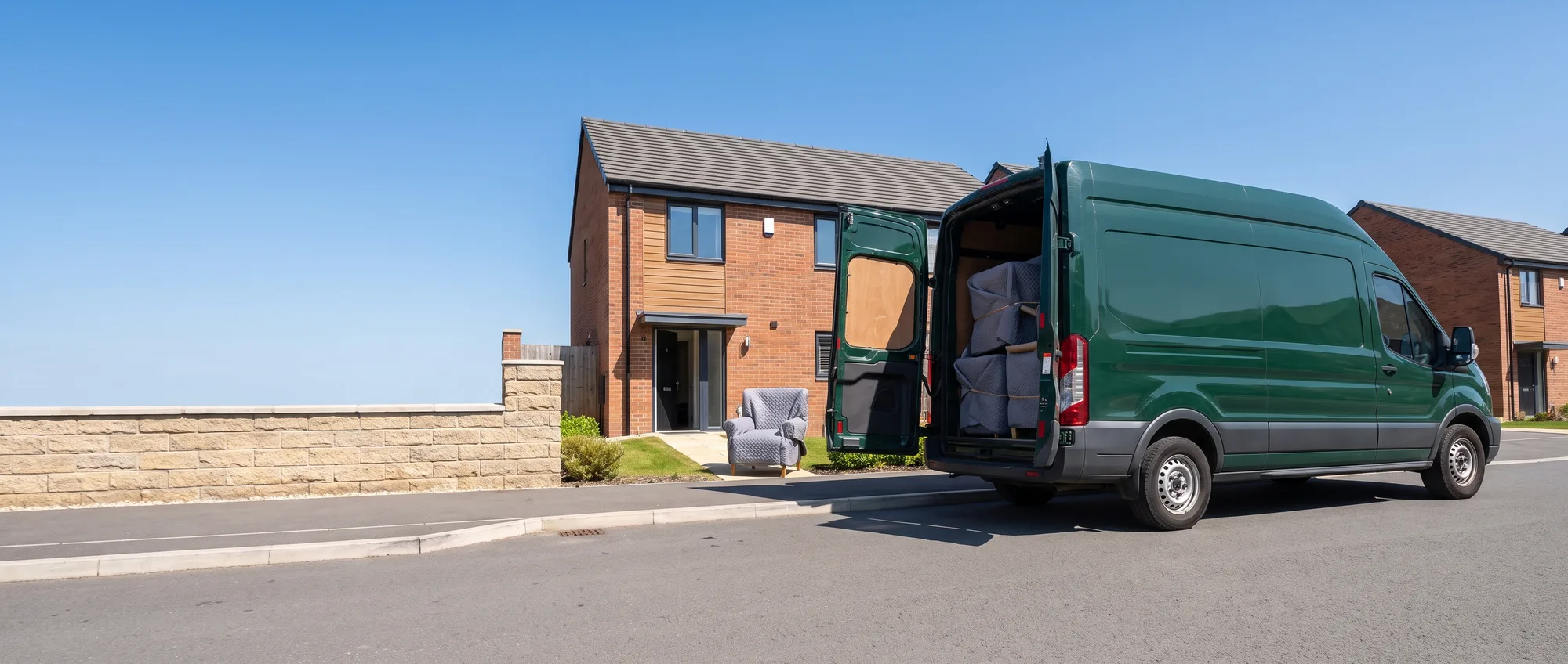 A green delivery van at the kerb outside a house on a bright day, its rear doors open on furniture wrapped in moving blankets, and an armchair set down on the path to the front door